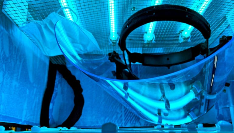 PPE (a face mask and plastic face shield) sit inside the UV disinfection box, with bright blue light illuminating its interior