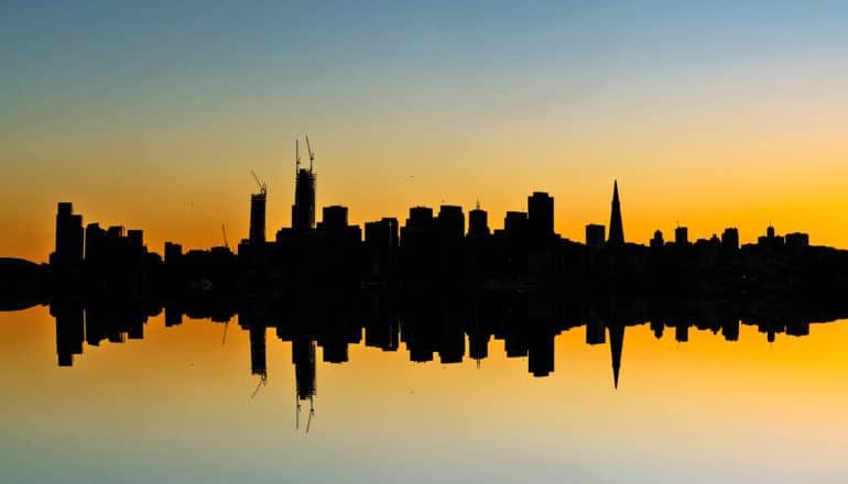 A city skyline at dusk reflects in water below, creating a perfect mirror of the skyline's silhouette