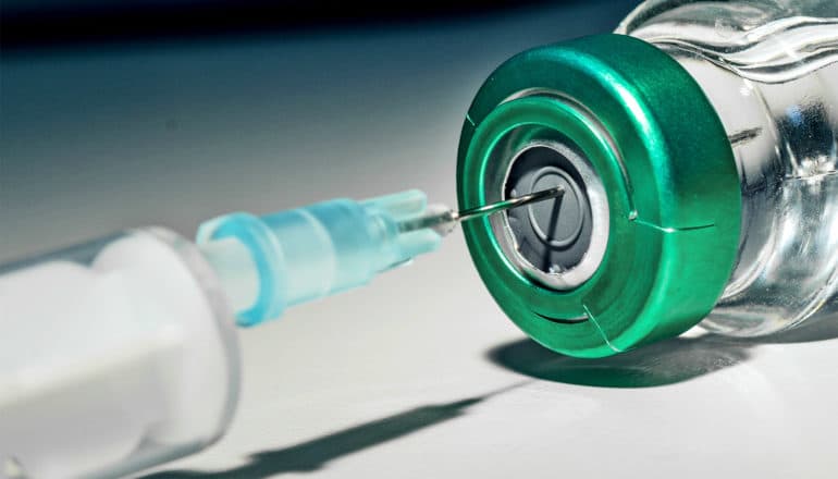 A syringe pulls liquid from a glass vaccine vial with a green top