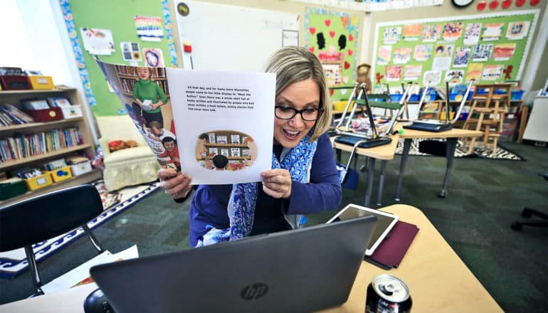 A teacher holds up a book to her laptop camera as she teaches remotely in an empty classroom