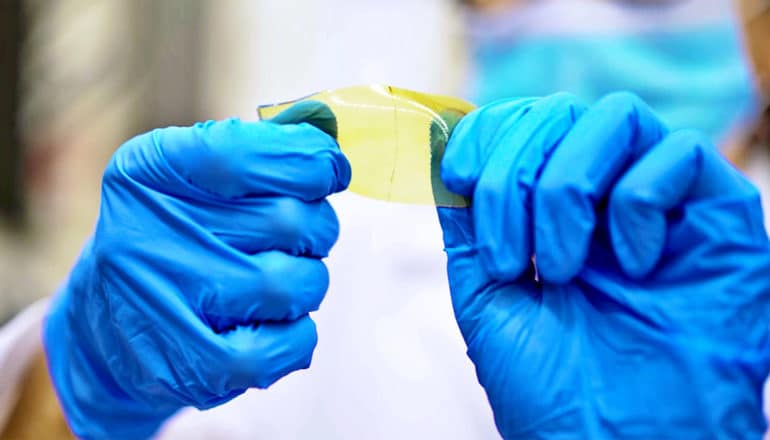 A researcher wearing blue gloves twists a thin, yellow piece of the self-healing synthetic polymer