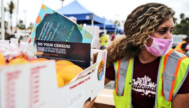 A woman in a mask hands out a crate of food containing census 2020 information at a food bank