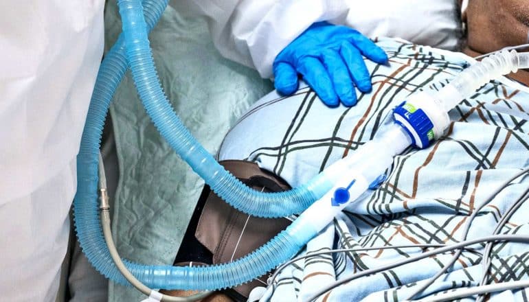 A health worker puts a blue-gloved hand on the shoulder of a patient on a ventilator