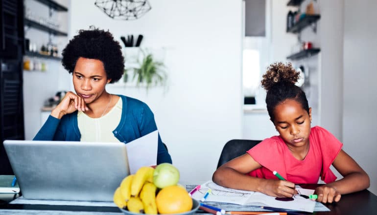 A mother on a laptop and daughter coloring both sit at the kitchen table