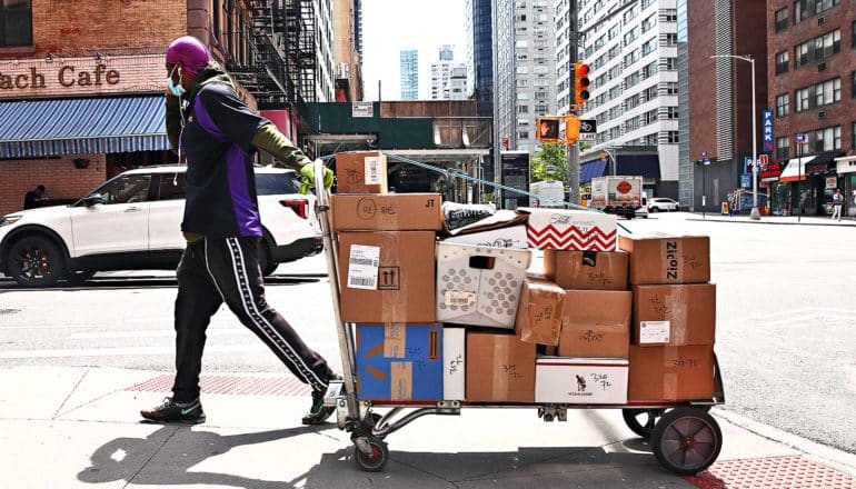 A man hauls a cart of packages on the sidewalk while wearing a mask