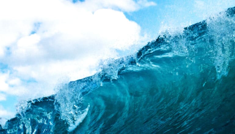 A deep blue ocean wave curls against a cloudy blue sky