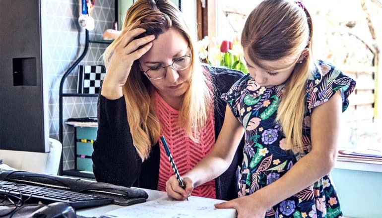 A mother looks down at her daughter's homework, holding her forehead as her daughter writes in a notebook