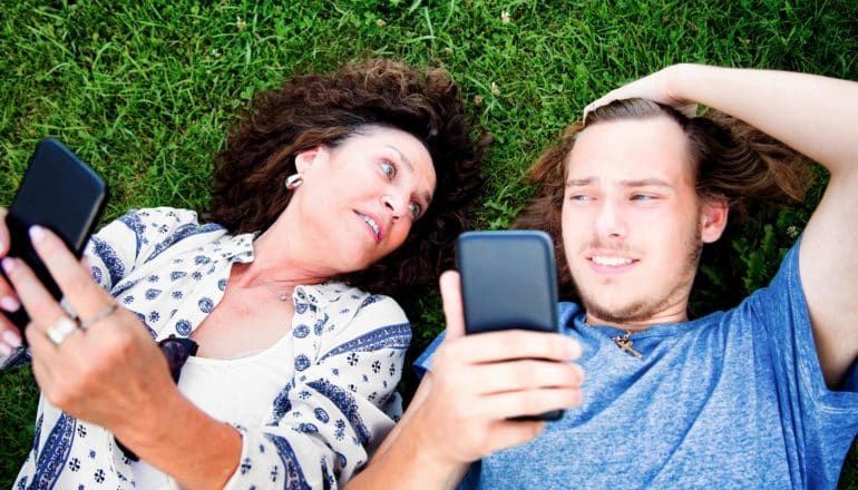 adult and young adult lie in grass with phones; older adult looks insistent, younger one looks exasperated