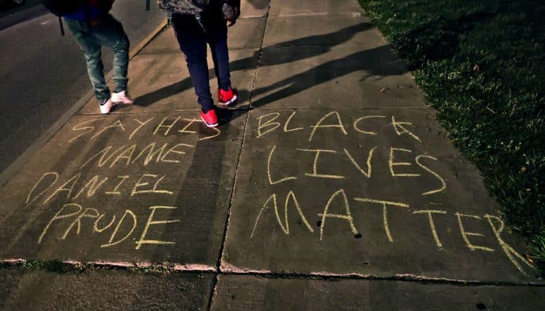 two people walk on sidewalk with chalk writing: "Say his name Daniel Prude. Black lives matter"