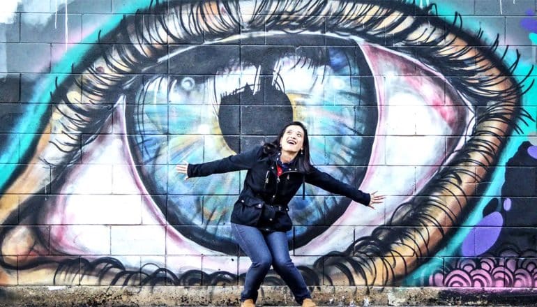 A young woman laughs and poses in front of a giant painting of an eyeball