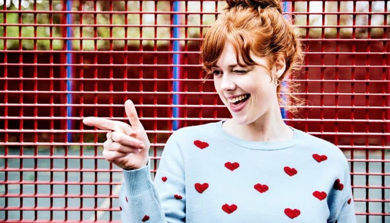 A woman in a heart-covered sweater winks and points off camera