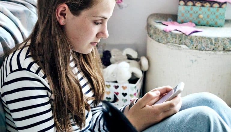 A teen looks down at her phone while sitting on her bedroom floor