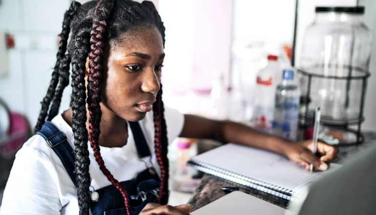 A young student looks at her laptop while taking notes, working on the kitchen counter