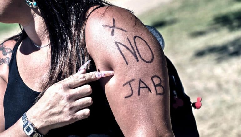 A woman points at her arm, where she's written "No Jab" and a large X