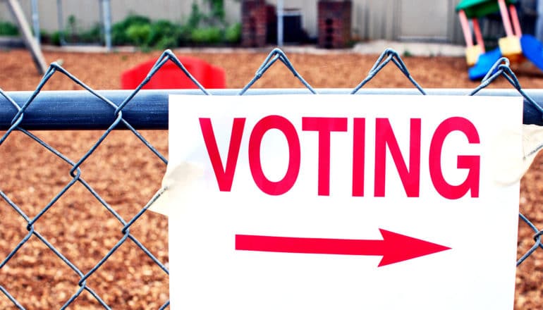 A sign on a chainlink fence outside a playground reads "Voting" and has an arrow pointing to the right