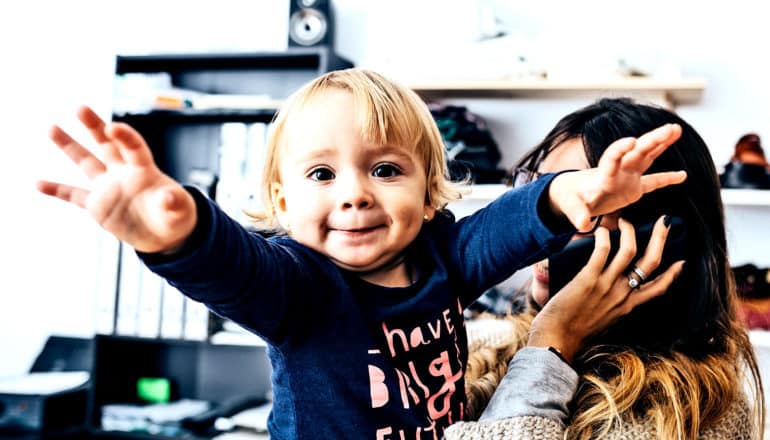 A baby girl reaches for the camera while her mother takes a work call