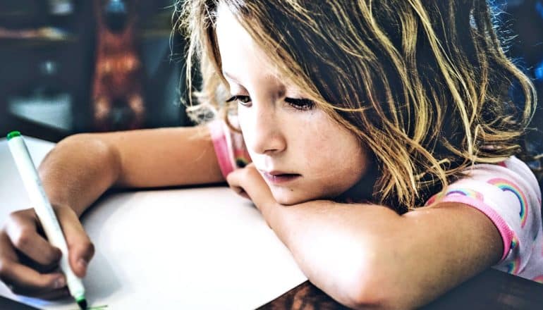 A young girl draws with a marker while leaning on her arm