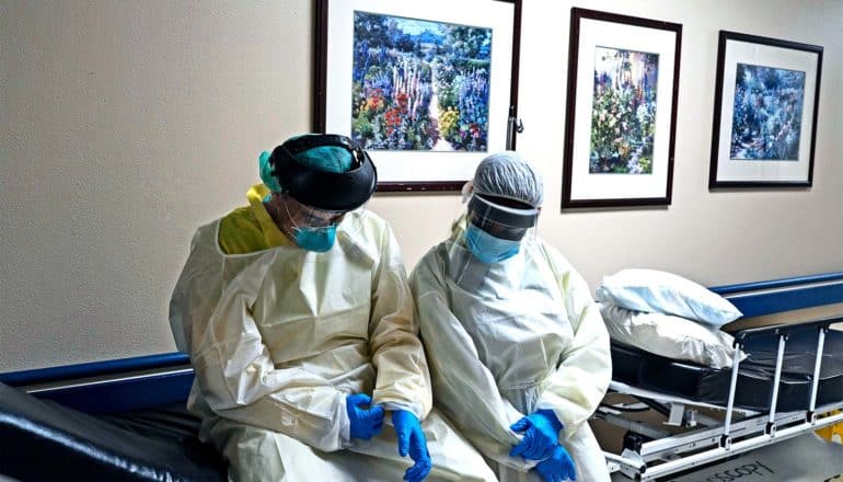 Two health workers in full protective gear sit on a stretcher in a hospital hallway looking down