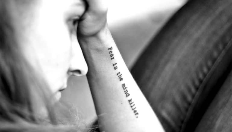 A young woman holds her forehead. A tattoo on her forearm reads, "Fear is the mind killer."