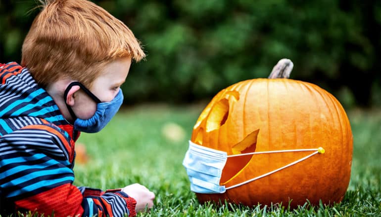 A young boy wearing a face mask looks at a Jack-o-lantern with a face mask