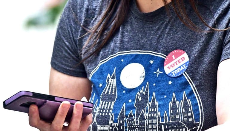 A young woman has an "I voted today!" sticker on her t-shirt and holds her iPhone