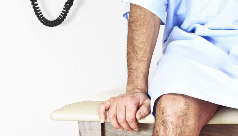 A man sits in a blue medical gown on an exam table at the doctor's office