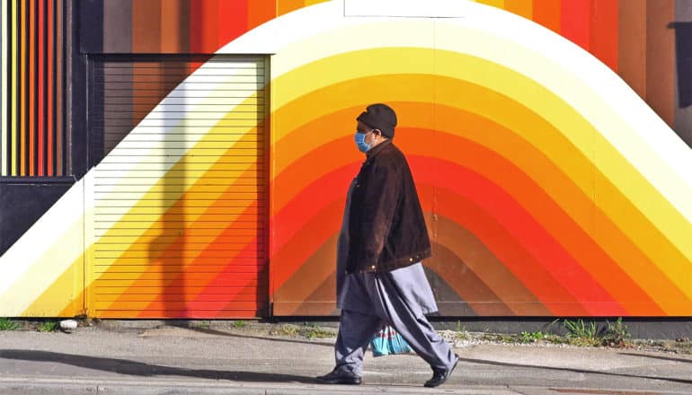 A man with a face mask walks past a multi-colored wall