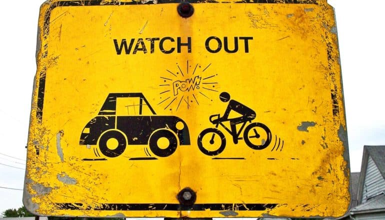 A yellow sign reads "Watch Out" and shows a car headed for a cyclist. Someone has written "Pow!" between the two