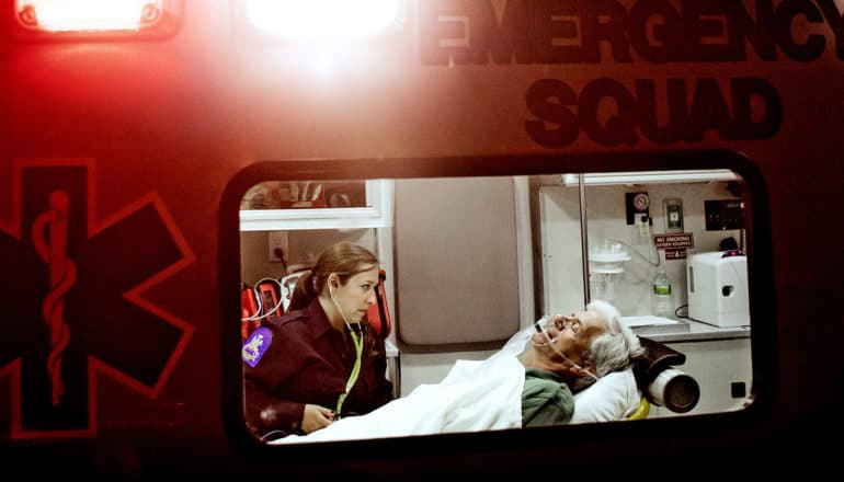 An EMS worker sits in the back of an ambulance with a patient on a stretcher