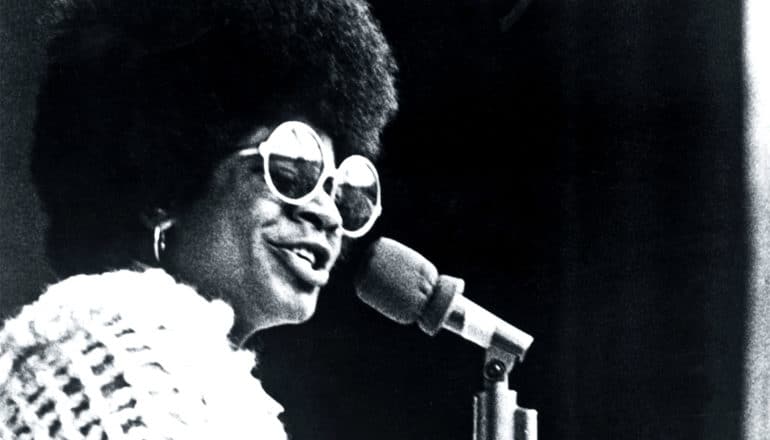 Merry Clayton sings into a microphone while wearing circular sunglasses