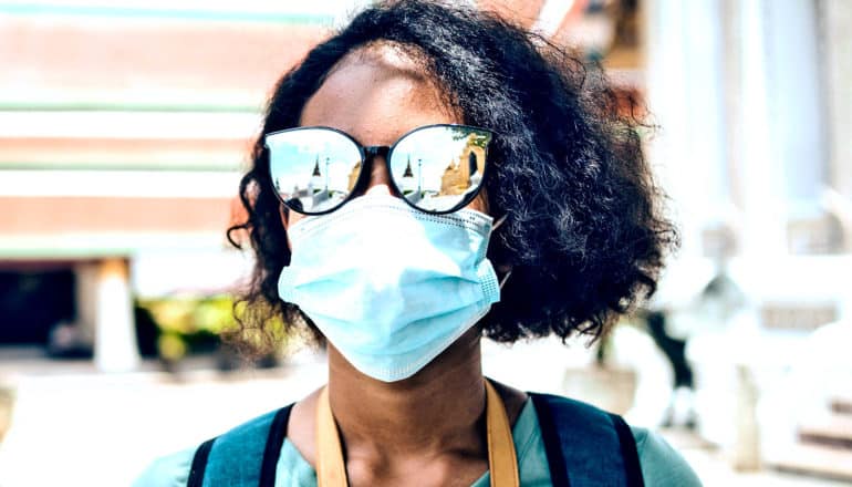 A college student in a mask and sunglasses walks with a backpack on