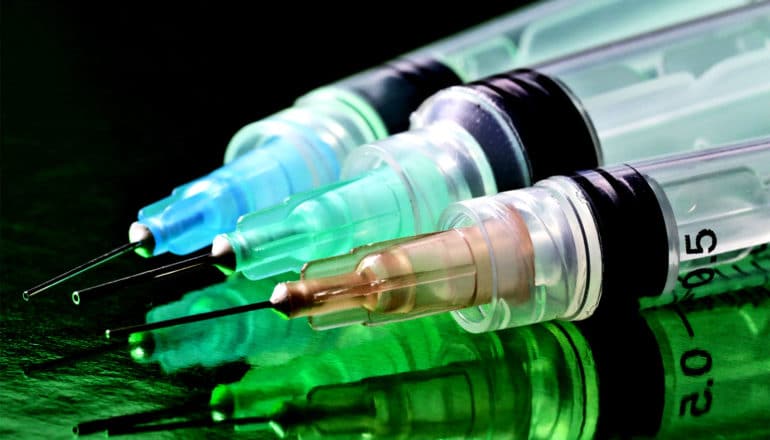 Three syringes sit on a reflective surface showing their reflection in green light