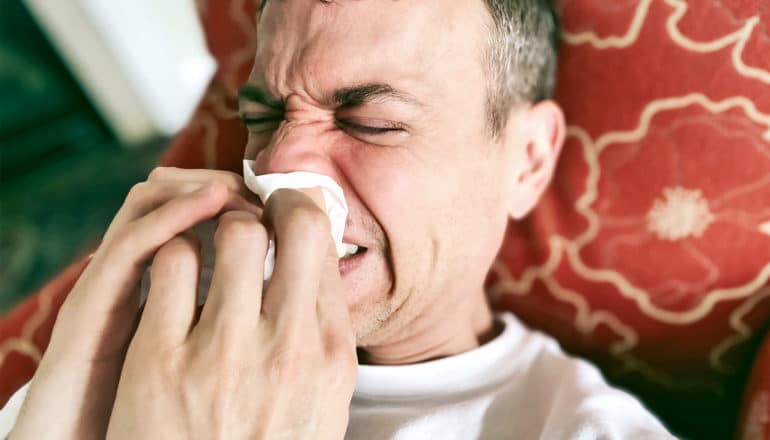 A man sneezes into a tissue while laying on a pillow on a couch