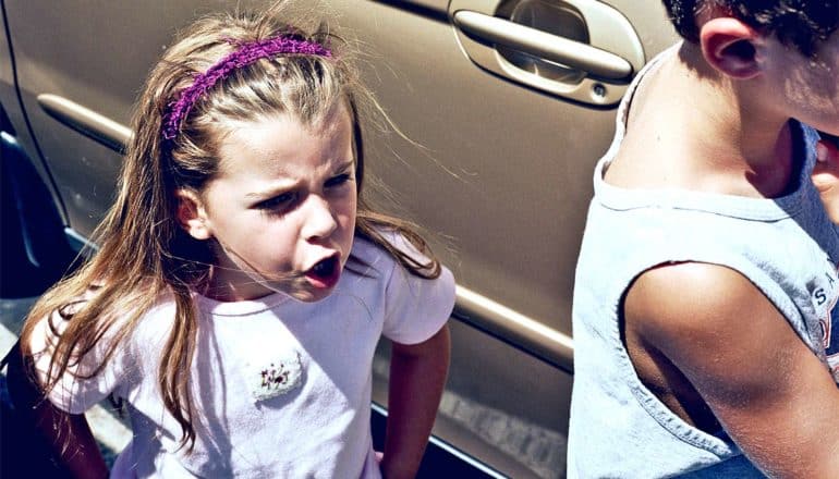 A young girl yells at a young boy as he walks away, with a car in the background