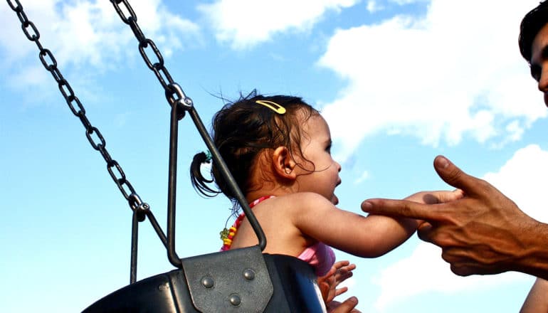 A child plays on a swing with her father nearby