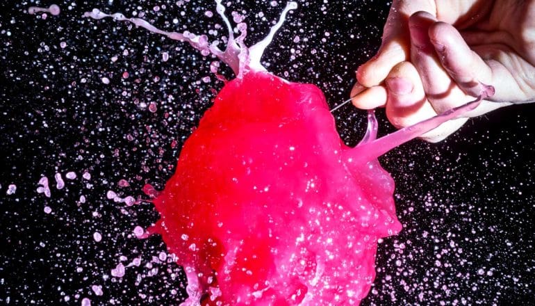 A person pops a red water balloon with a pin as water shoots everywhere against a black background