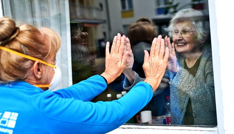 A woman in a face mask and an older woman inside a nursing home touch hands through the glass