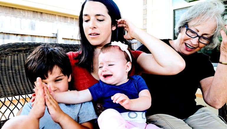 A mother holds a baby who is crying and reaching out to her brother, with a grandmother looking on