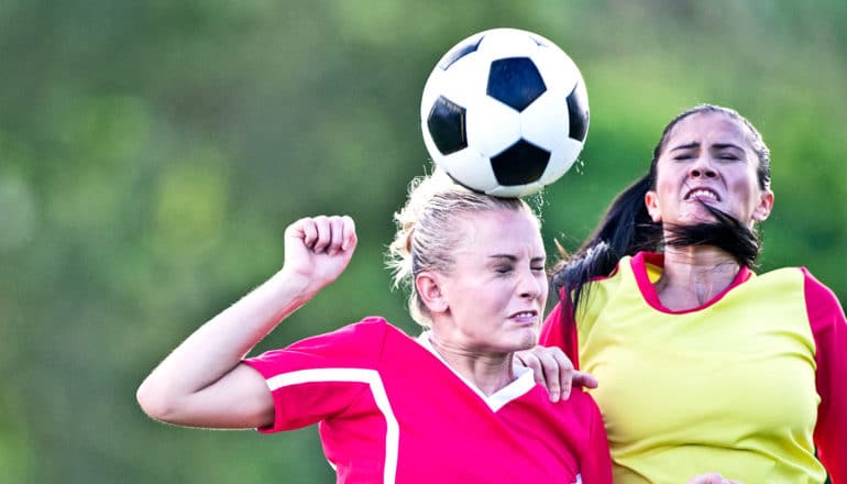 Two soccer players jump for a header, both grimacing and closing their eyes