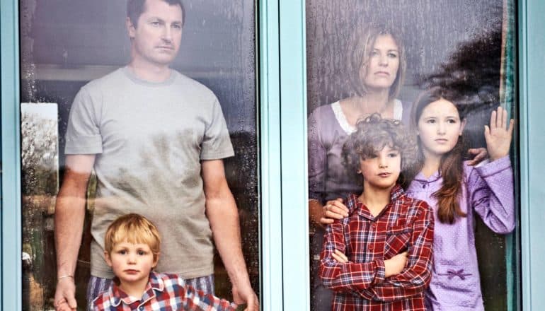 A family stands inside looking out the window with blank faces