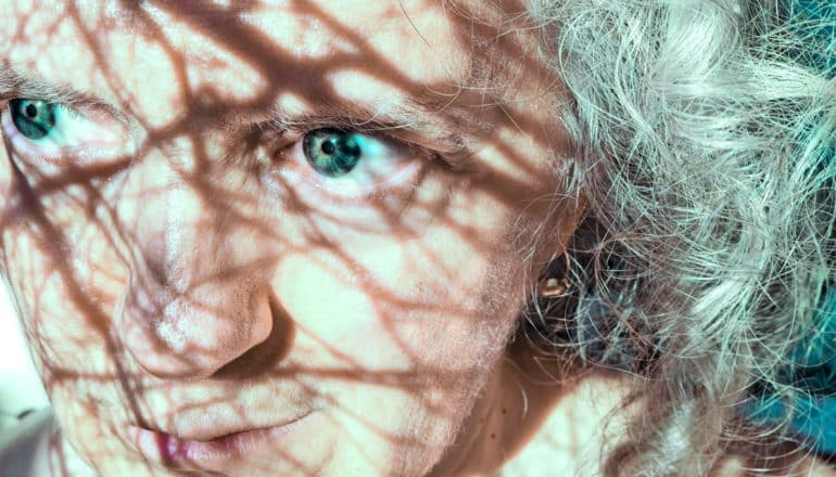 An older woman looks up as shadows from tree branches fall on her face