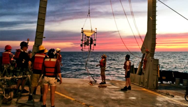 Researchers stand on the deck of a ship while lowering the device by winch into the water. The sun sets in the background.