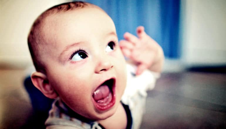 A baby holds his hand up to his ear and looks up as if he's listening intently