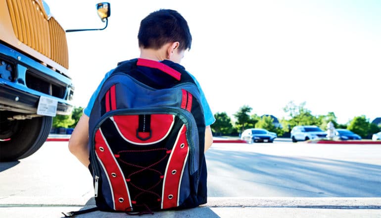 A young boy sits alone on the curb near a school bus and parking lot