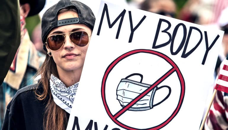 A young woman holds a sign that reads "My Body, My Choice" with a picture of a mask with a "no" symbol over it