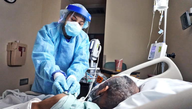 A health worker in full protective gear gets info about a patient in a hospital bed