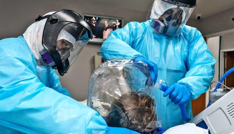 Two health workers in full protective gear place a tube into the plastic helmet ventilator