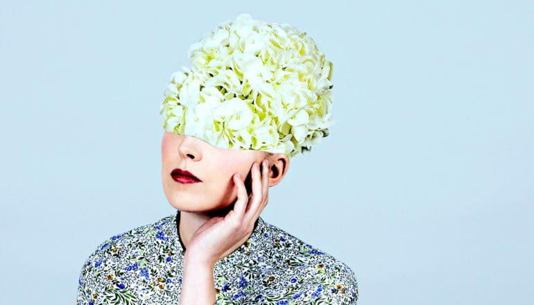 A woman puts her hand to her face and looks lost in thought as flowers cover her eyes and the top of her head