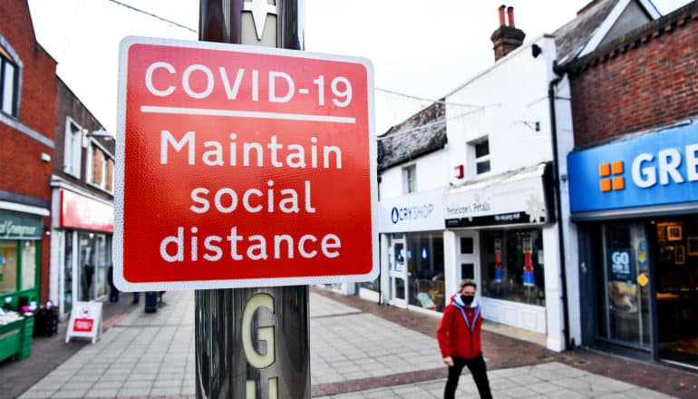 A red sign reads "COVID-19 Maintain social distance" and a man walks past wearing a face mask