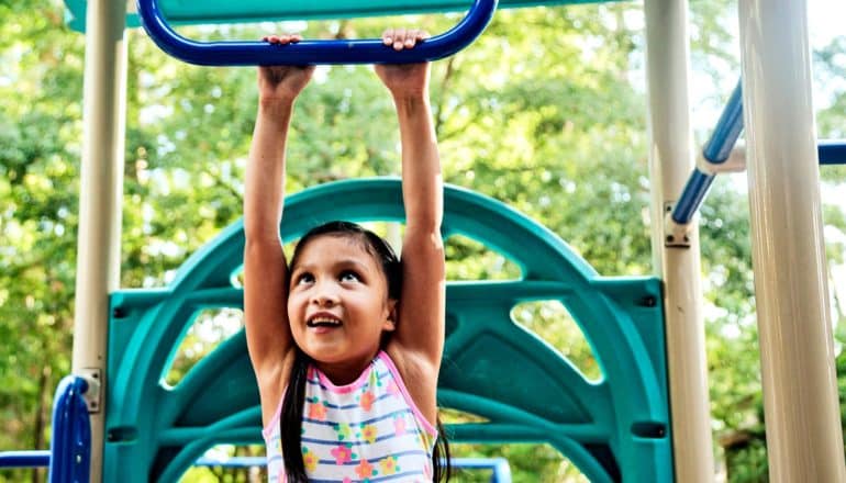 A young girl hangs from monkey bars at a park